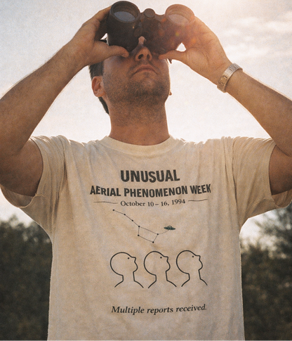 Man wearing a t-shirt with text and graphics, looking through binoculars outdoors.