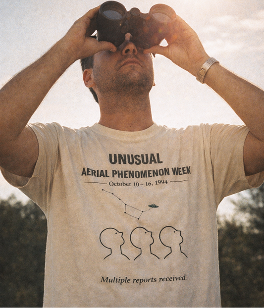 Man wearing a t-shirt with text and graphics, looking through binoculars outdoors.